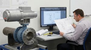 An engineer reviews a butterfly valve drawing in an industrial piping project evaluation area, with an actuated butterfly valve assembly in the foreground and dimensional, interface, and revision details displayed on a monitor and printed drawings.
